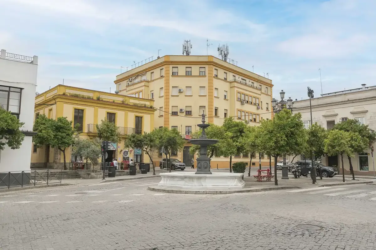 A paved city square with a central fountain and trees in front of surrounding buildings.