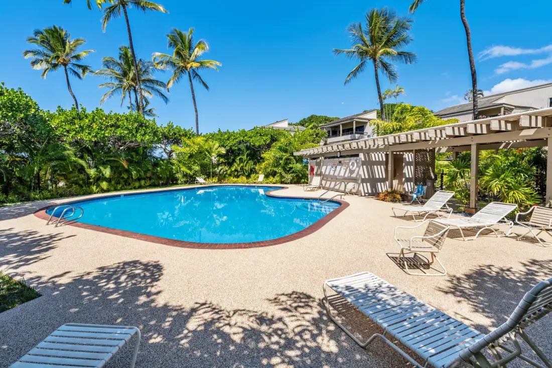 An outdoor swimming pool sits beside a shaded seating area and tropical greenery.