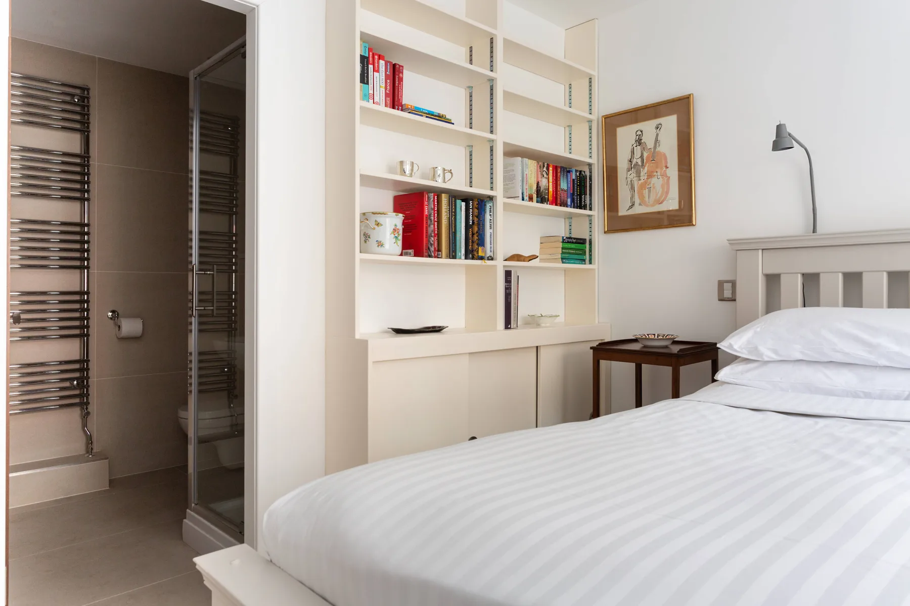 A bedroom with a neatly made bed beside built-in bookshelves and an open shower area.