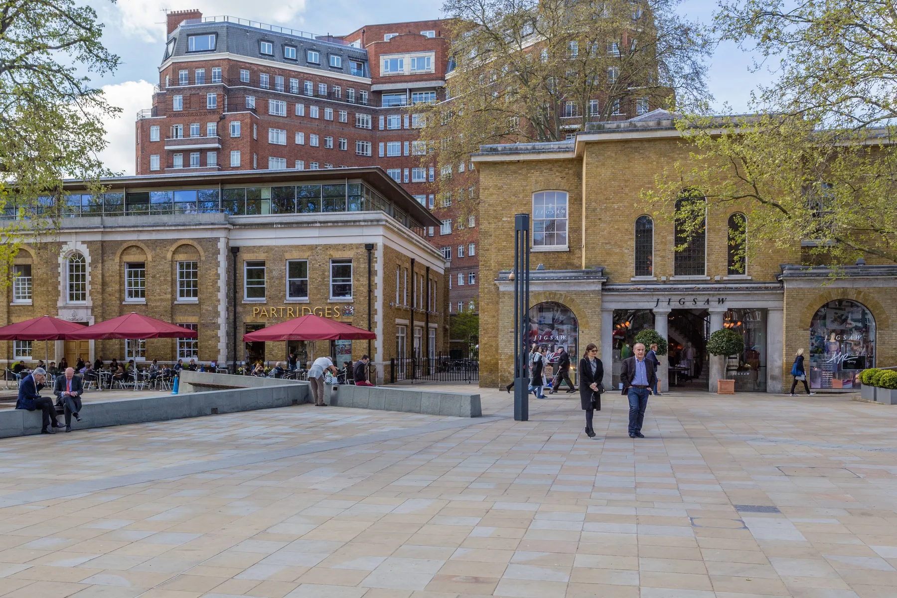 A paved outdoor plaza fronts brick buildings with café seating and trees.