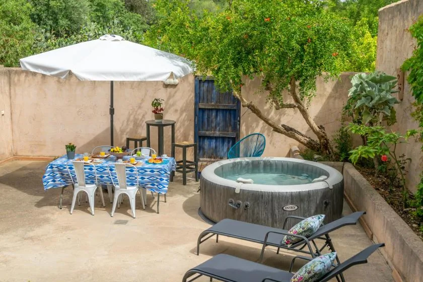 An outdoor patio has a dining table, a hot tub, and lounge seating amid greenery.