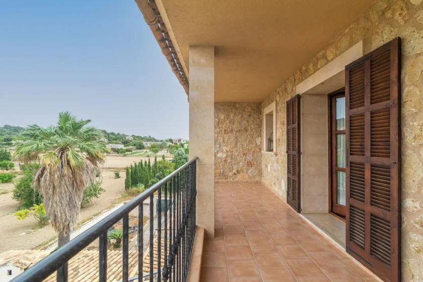 A covered balcony with tiled flooring and shuttered doors overlooks a green landscape.