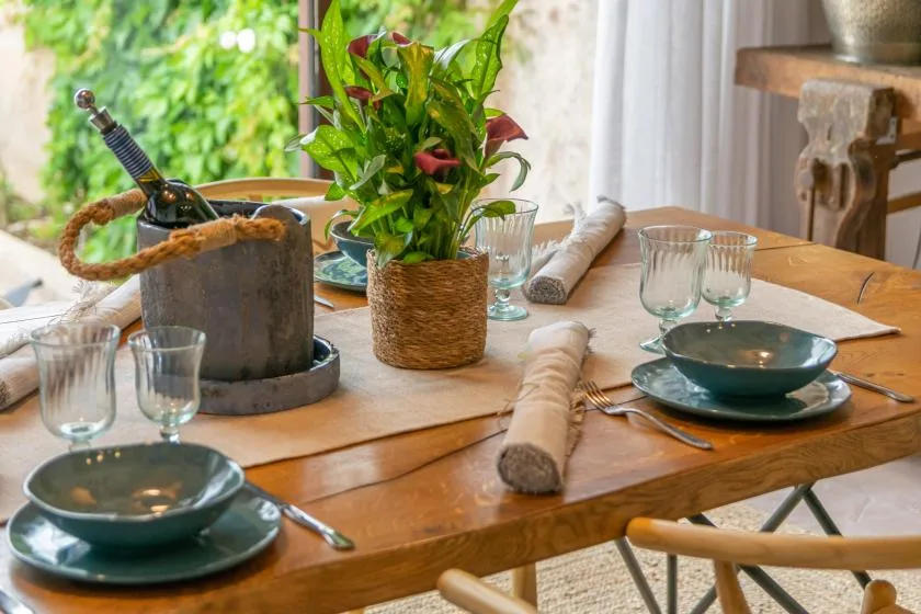 A dining table is set with dishes, glassware, rolled napkins, and a leafy centerpiece.