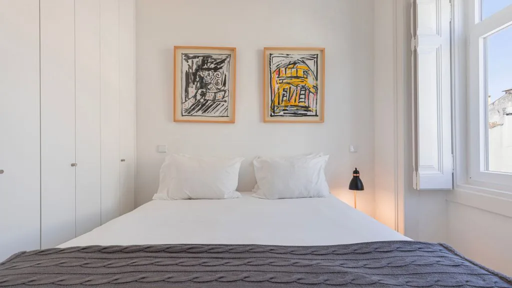 Un dormitorio luminoso con una cama bien tendida y cuadros enmarcados encima.