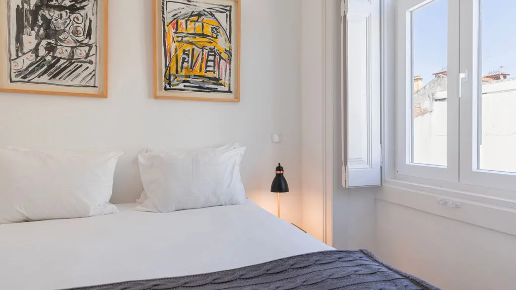 Un dormitorio luminoso con una cama bien tendida y cuadros enmarcados encima.