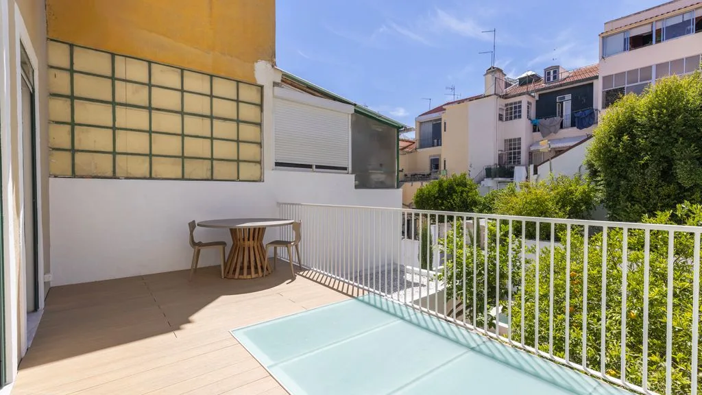Una terraza exterior con suelo de baldosas tiene una mesa pequeña y asientos junto a una barandilla blanca.