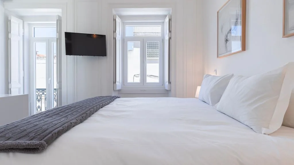 Un dormitorio luminoso con ropa de cama blanca y ventanas altas.