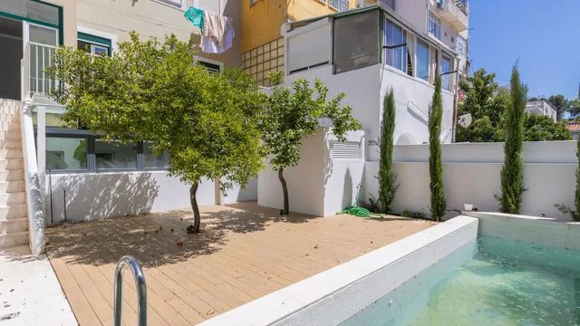 Una piscina junto a un patio pavimentado y árboles frondosos.