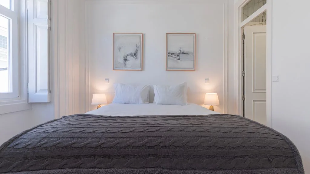 Un dormitorio luminoso con una cama tendida y cuadros enmarcados sobre ella.