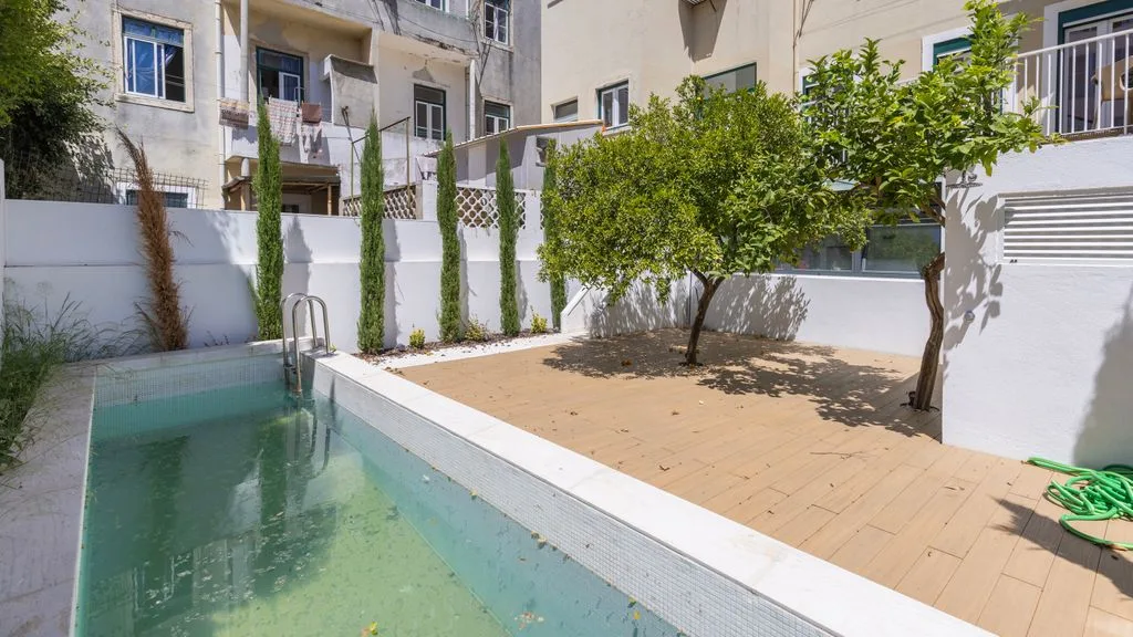 Una piscina en un patio junto a una terraza pavimentada, árboles y edificios alrededor.