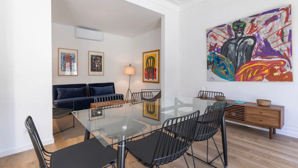 Un comedor con mesa de vidrio y sillas negras de alambre está junto a una sala de estar.