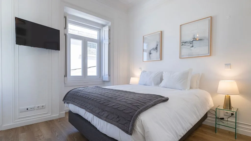 Un dormitorio luminoso con cama doble, televisor en la pared y cuadros.
