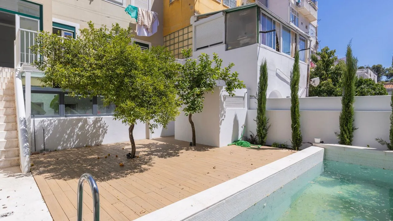 Una piscina al aire libre junto a un patio de baldosas, árboles y un muro blanco.