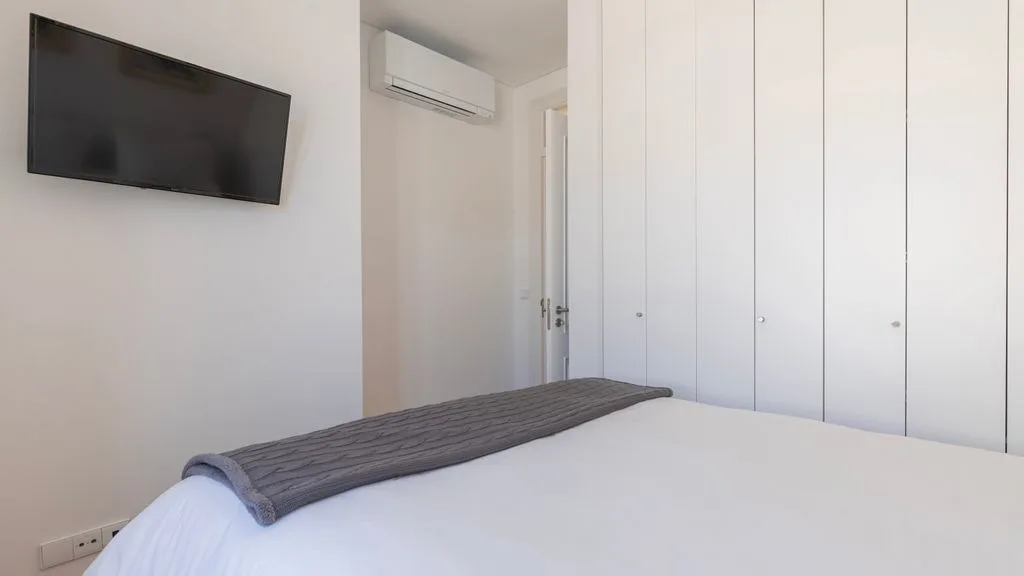 Un dormitorio luminoso con una cama bien tendida y un televisor en la pared.