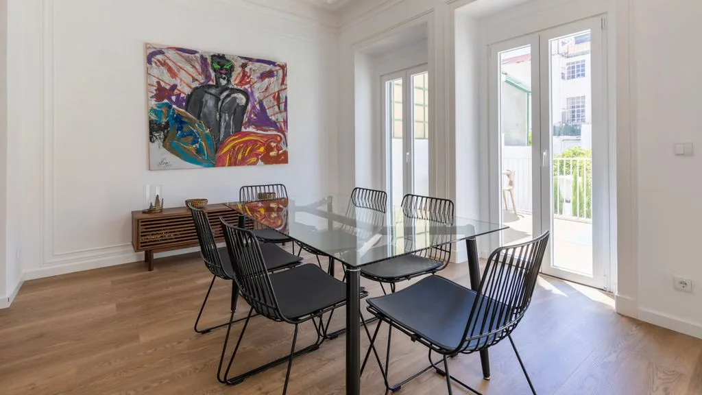 Un comedor luminoso tiene una mesa de vidrio y una obra de arte colorida en la pared.