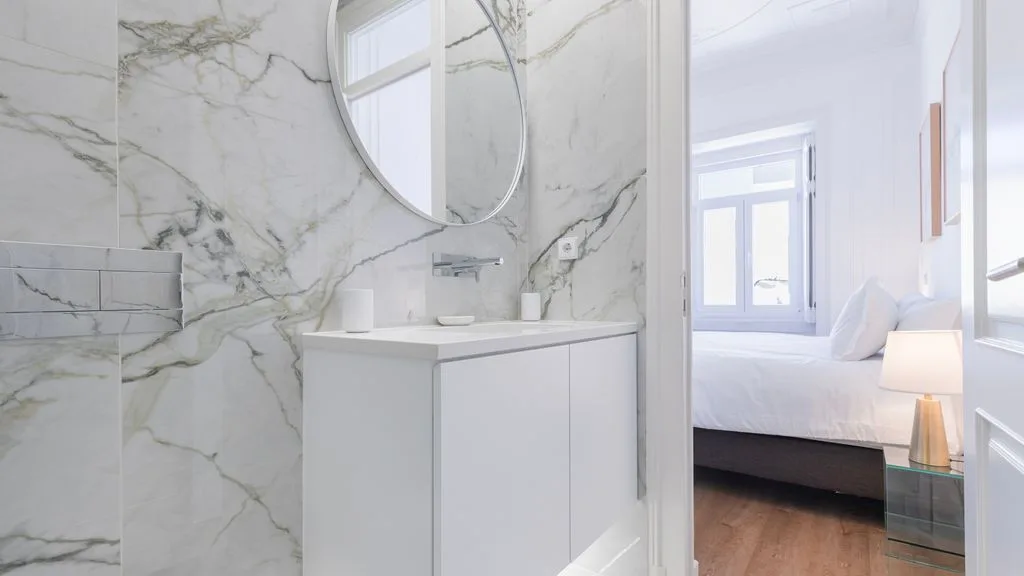 Un lavabo de baño con acabado de mármol y espejo redondo da a un dormitorio.