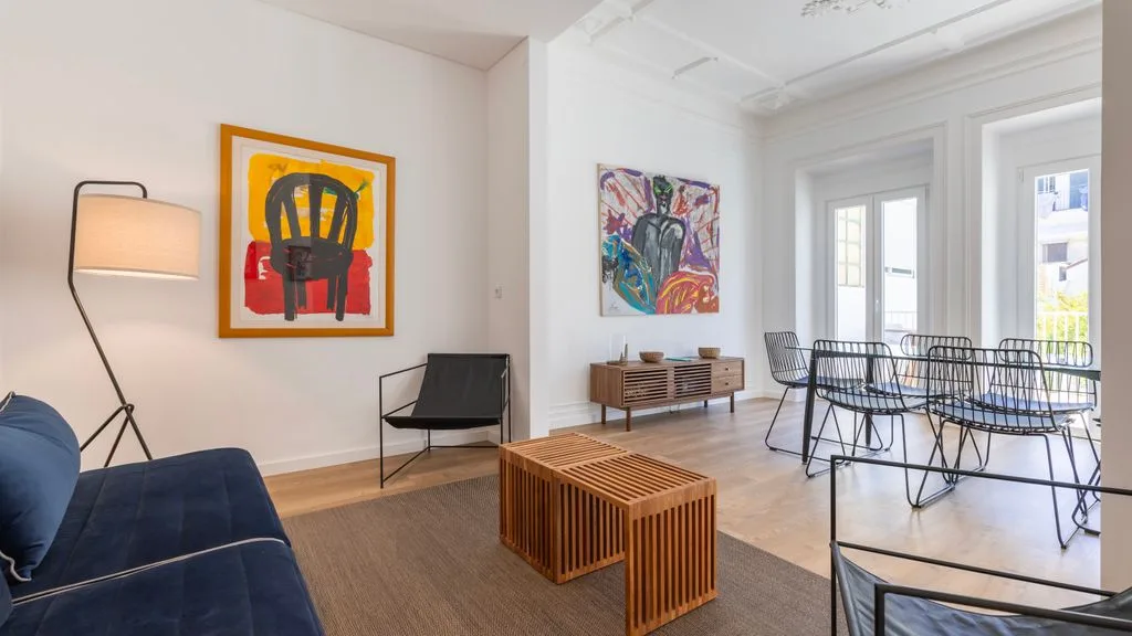 Un salón luminoso con asientos, cuadros coloridos y una mesa de comedor junto a las ventanas.