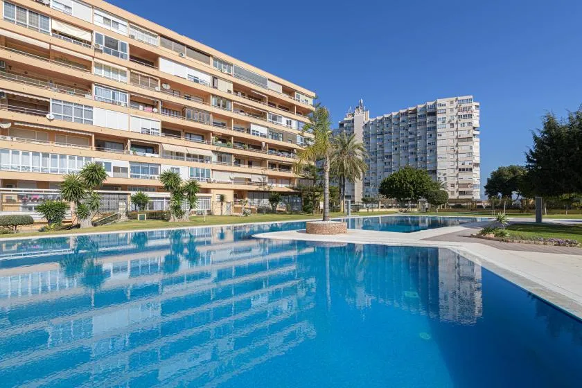 A large outdoor swimming pool reflects nearby apartment buildings beneath a clear blue sky.