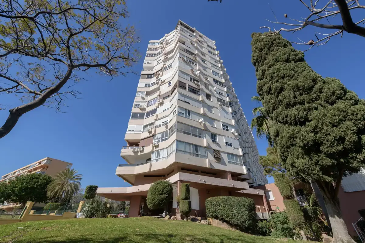 A tall apartment building stands above a landscaped lawn beneath a clear blue sky.