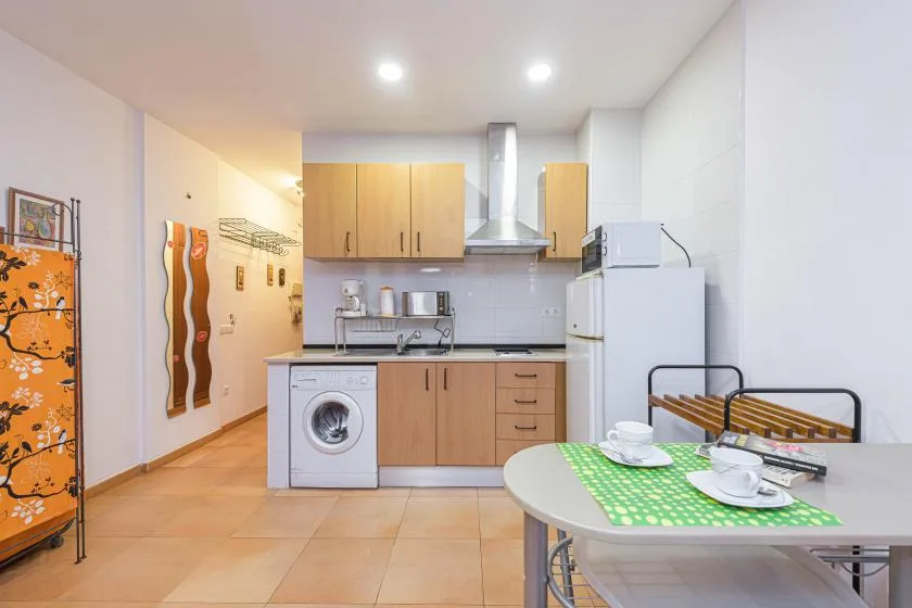 A compact kitchen with light wood cabinets, a washing machine, and a dining table.