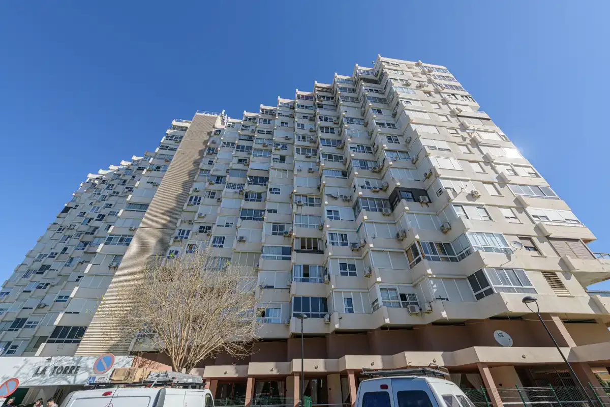 A tall apartment building rises above storefronts under a clear blue sky.