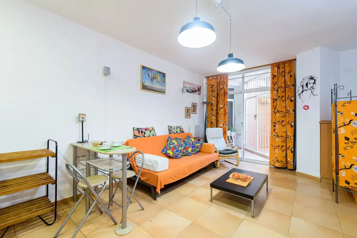 A bright living room with orange seating, a small dining table, and balcony doors.