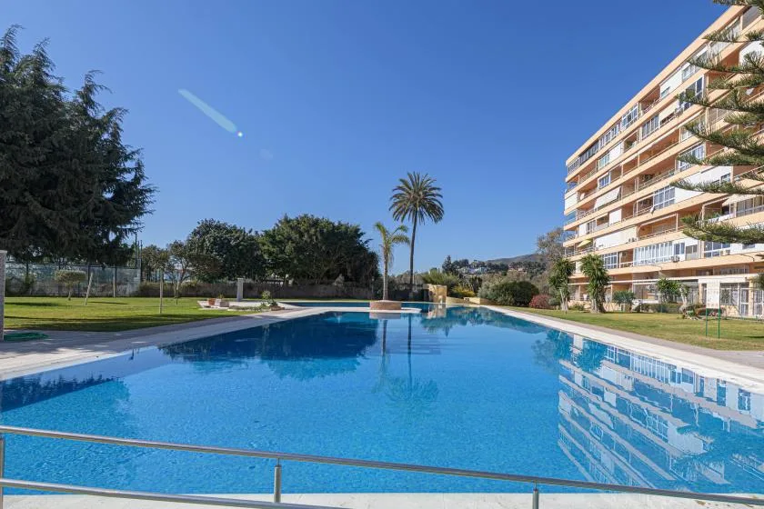 An outdoor swimming pool reflects a nearby apartment building under a clear blue sky.