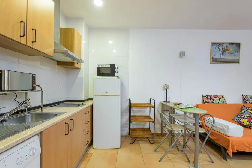 A compact kitchen and dining area with a table beside an orange-covered bed.