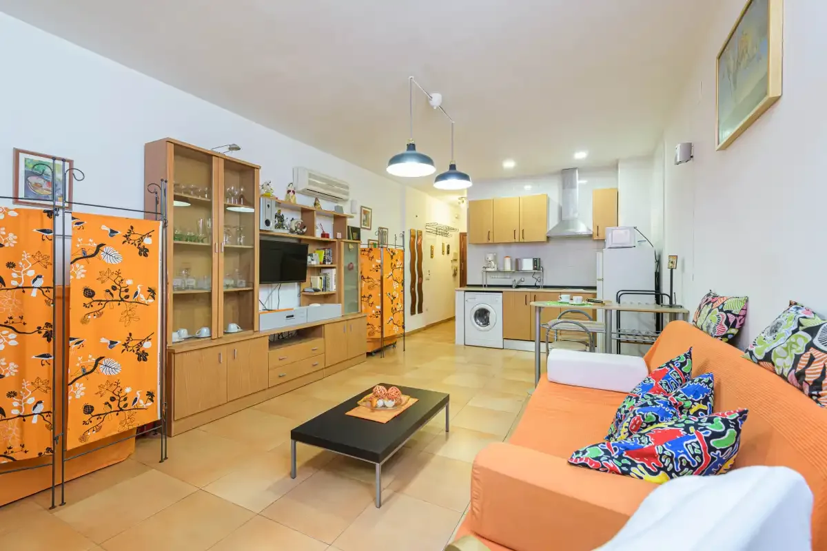 A bright studio living area with orange seating, a coffee table, and a compact kitchen.