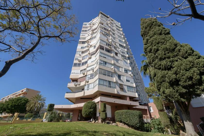 A tall residential building rises above a landscaped lawn under a clear blue sky.