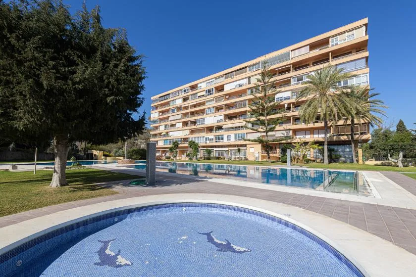 Outdoor swimming pools sit in front of a multistory building under a clear blue sky.