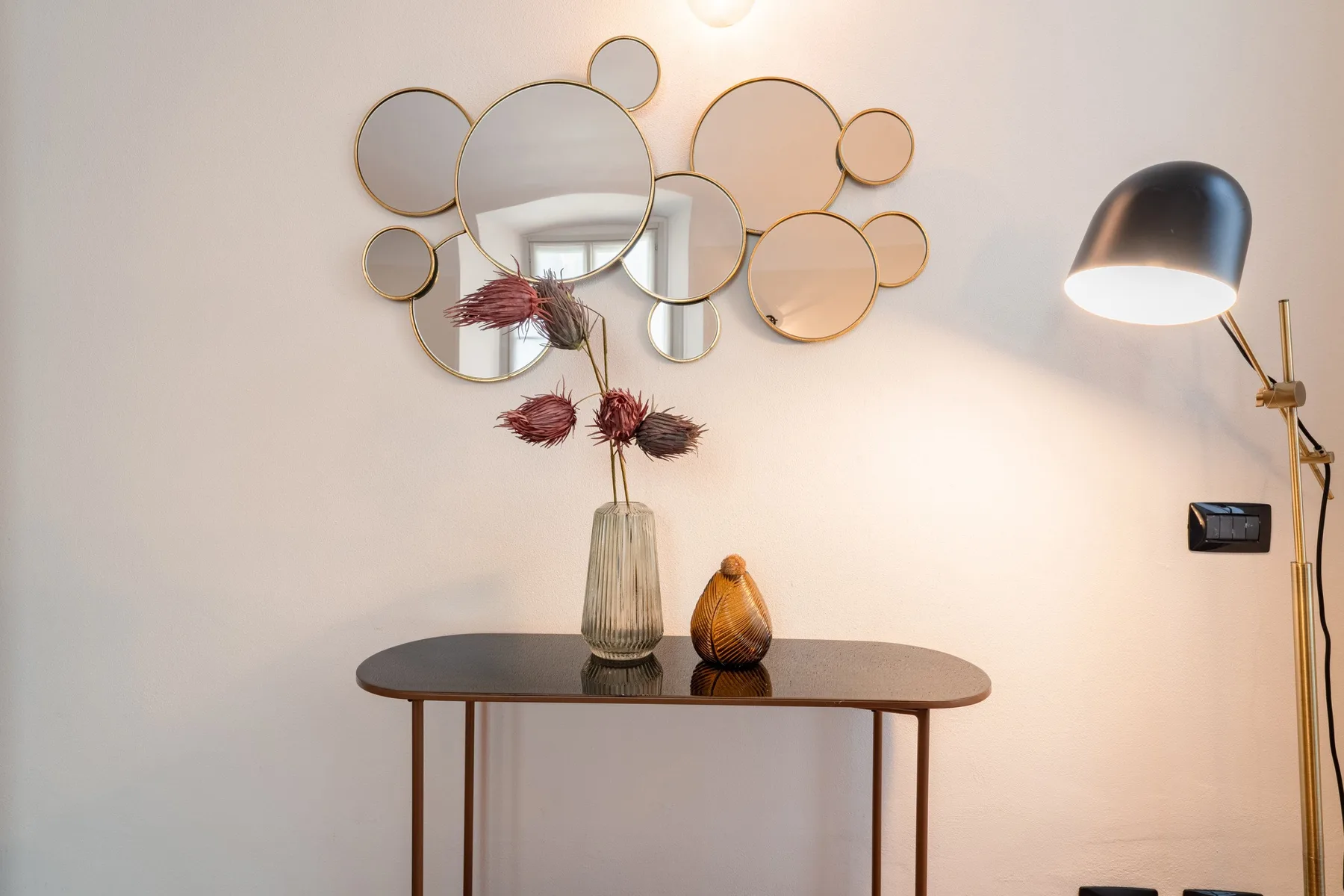 Una mesa consola con espejos decorativos y flores secas está junto a una pared clara.