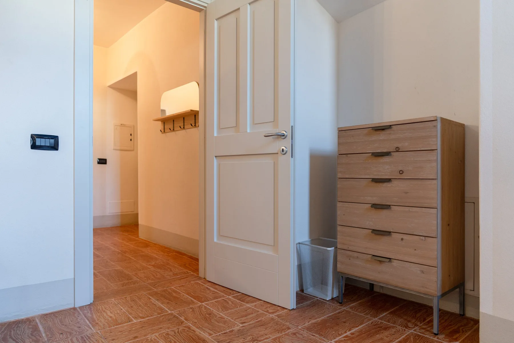 Una habitación luminosa con una puerta abierta, una cómoda de madera y una papelera pequeña.