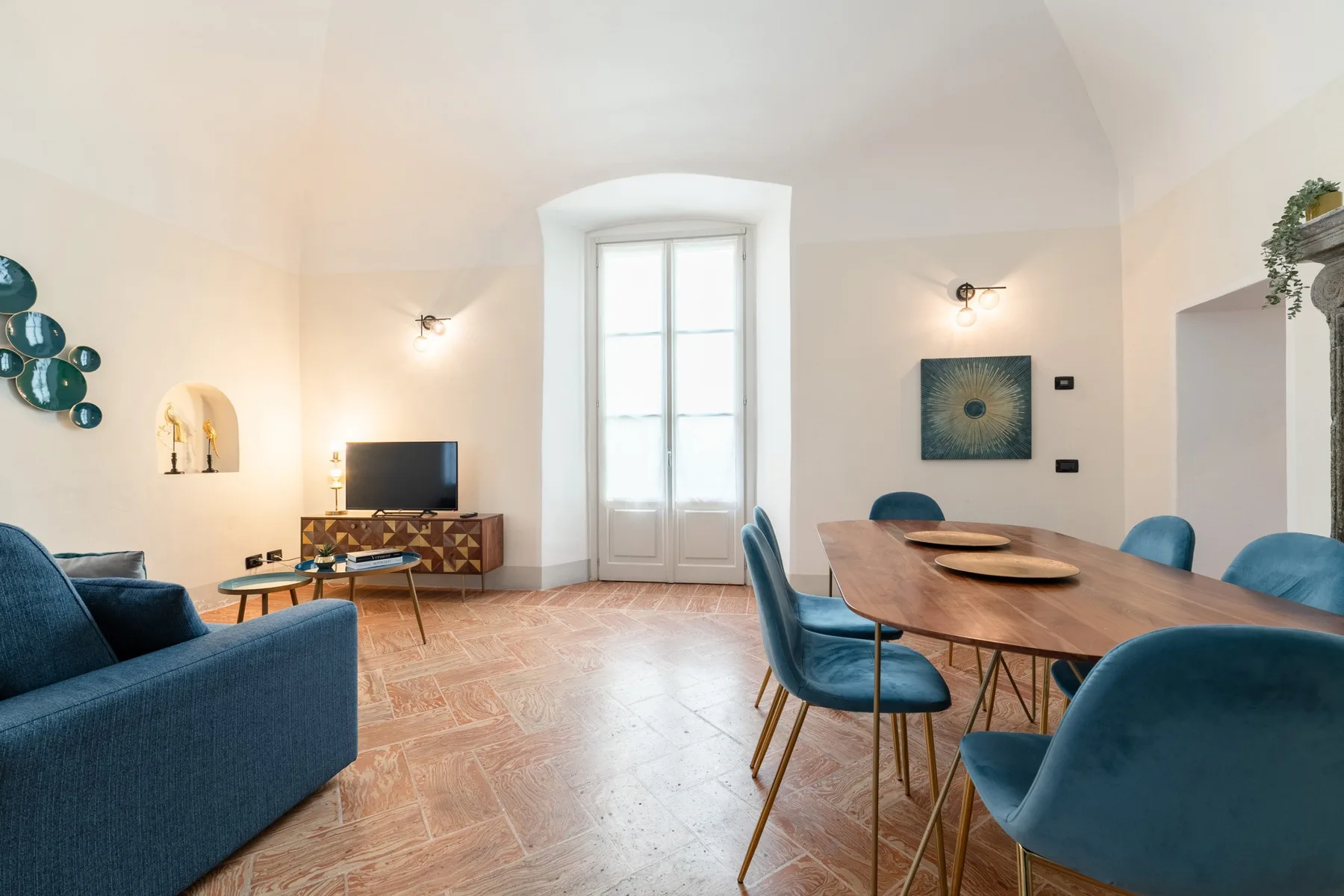 Una luminosa sala de estar y comedor con asientos azules, una mesa de madera y ventanas altas.