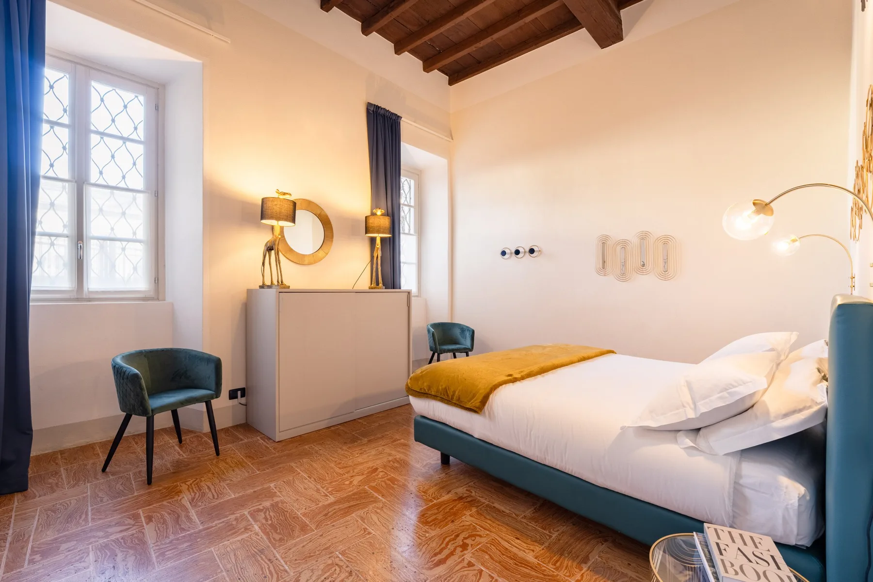 Un dormitorio con una cama, dos sillas y ventanas altas.