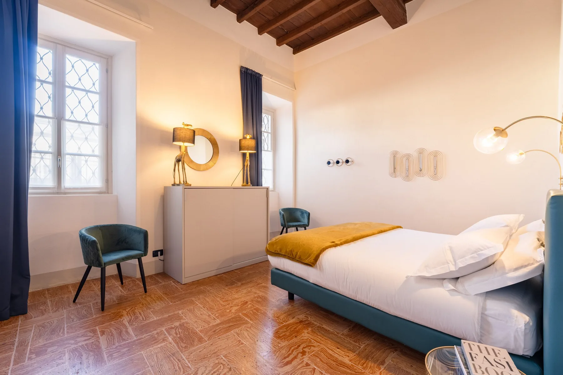 Un dormitorio con una cama, un escritorio pequeño y ventanas altas.