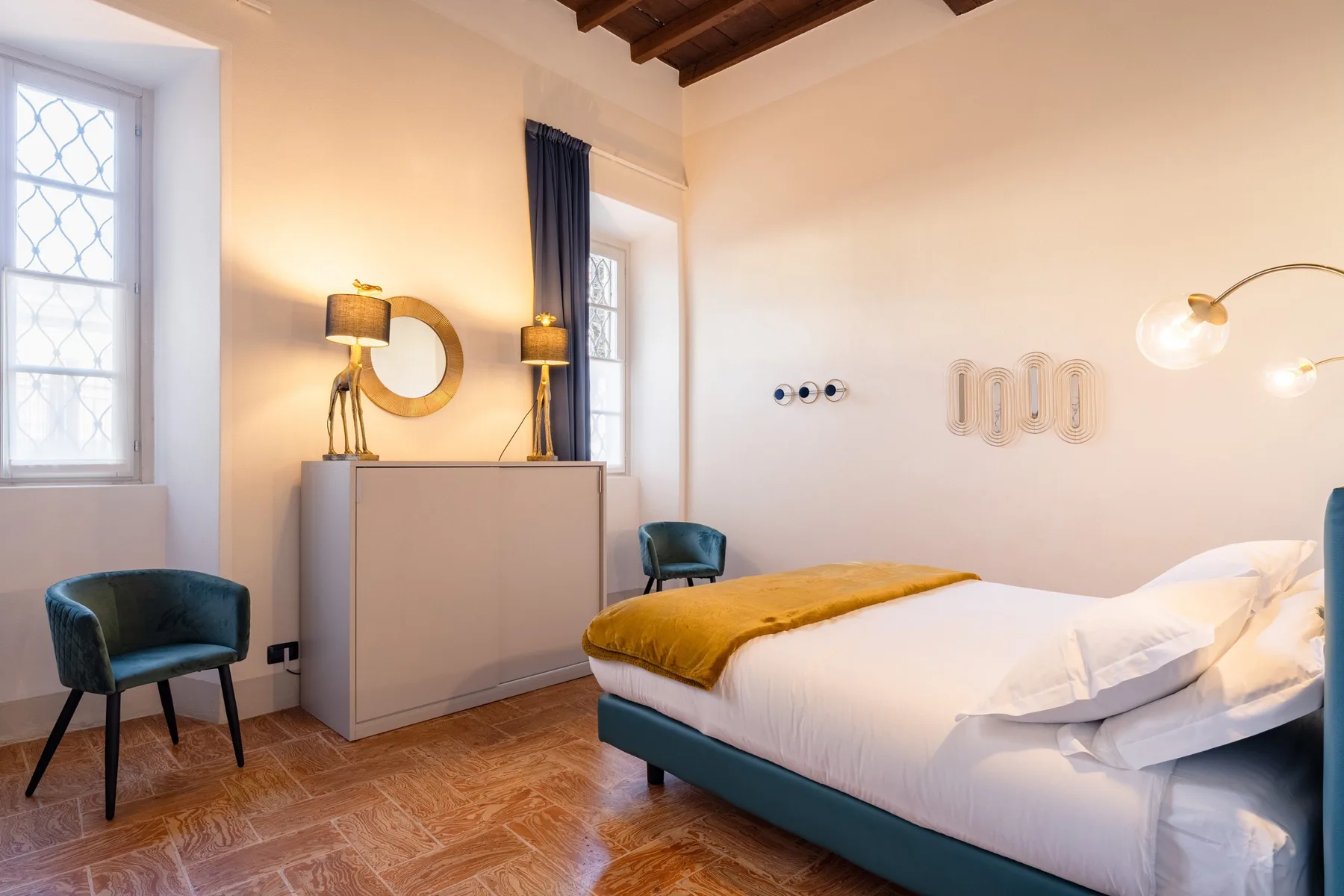 Un dormitorio con una cama, asientos verde azulado y un mueble bajo las ventanas.