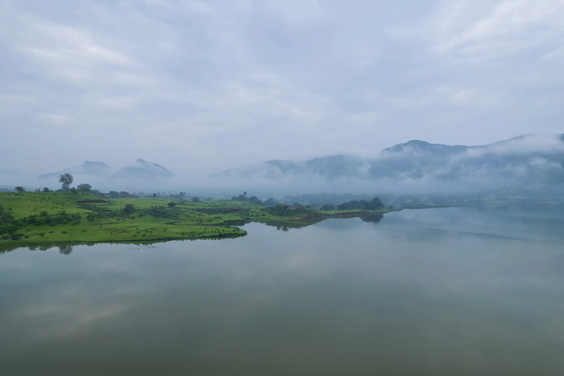 A calm lake reflects misty mountains and green hills beneath a cloudy sky.