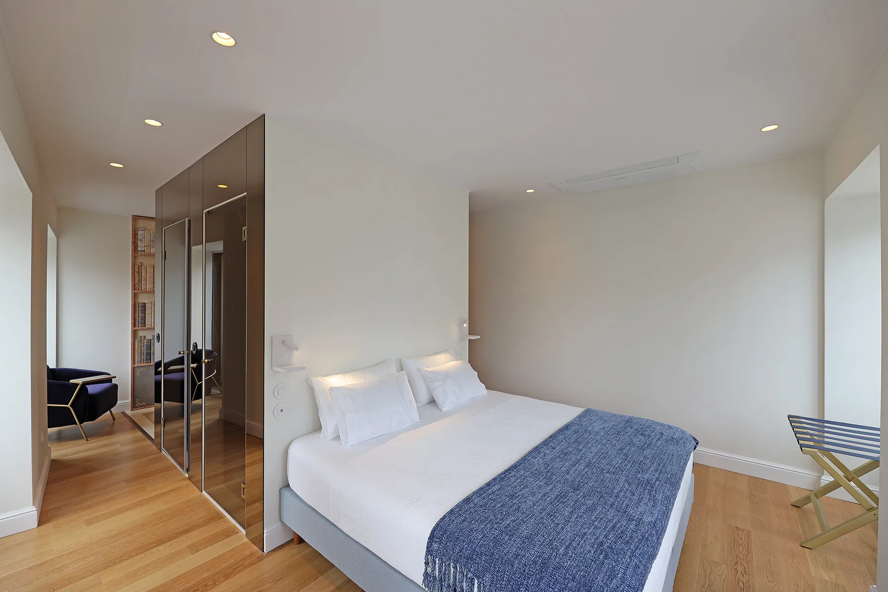 Un dormitorio luminoso con una cama blanca y una colcha azul junto a armarios con espejo.