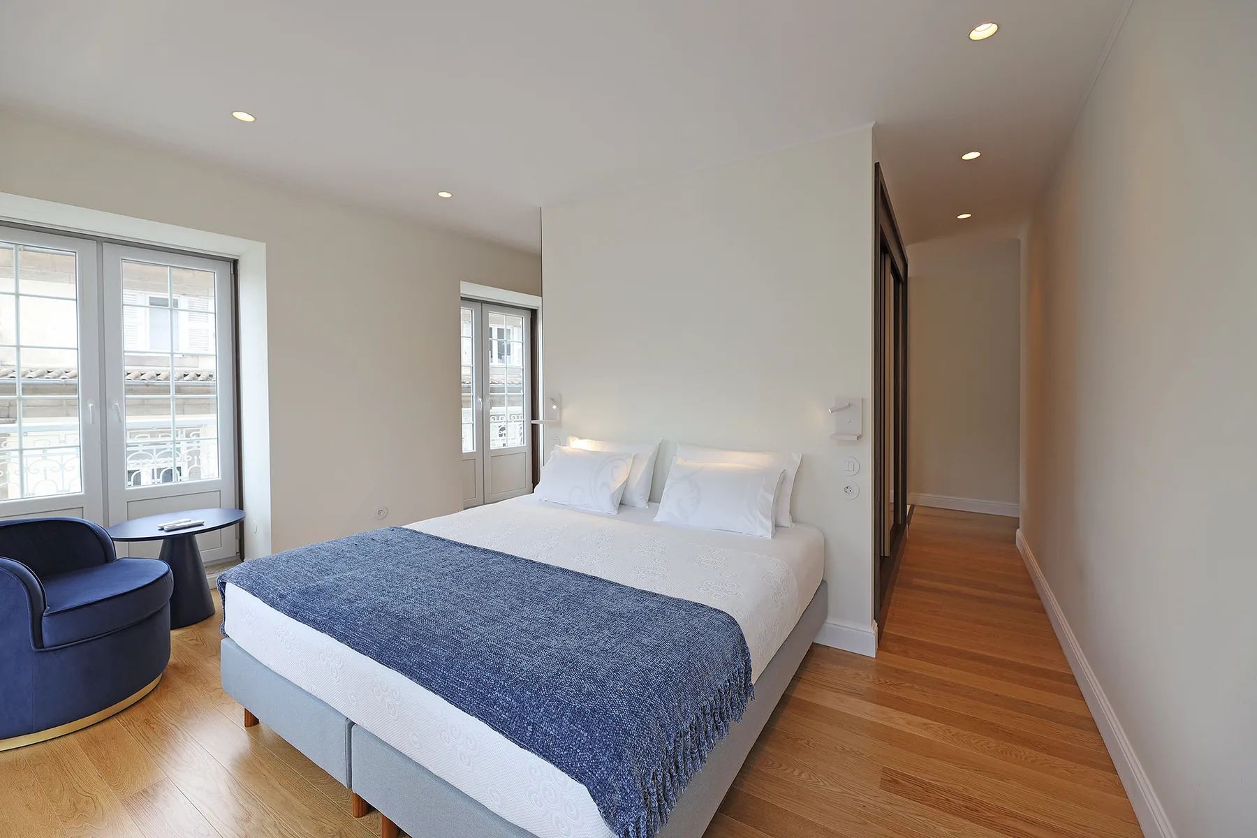 A bright bedroom with a double bed, blue blanket, and large windows.