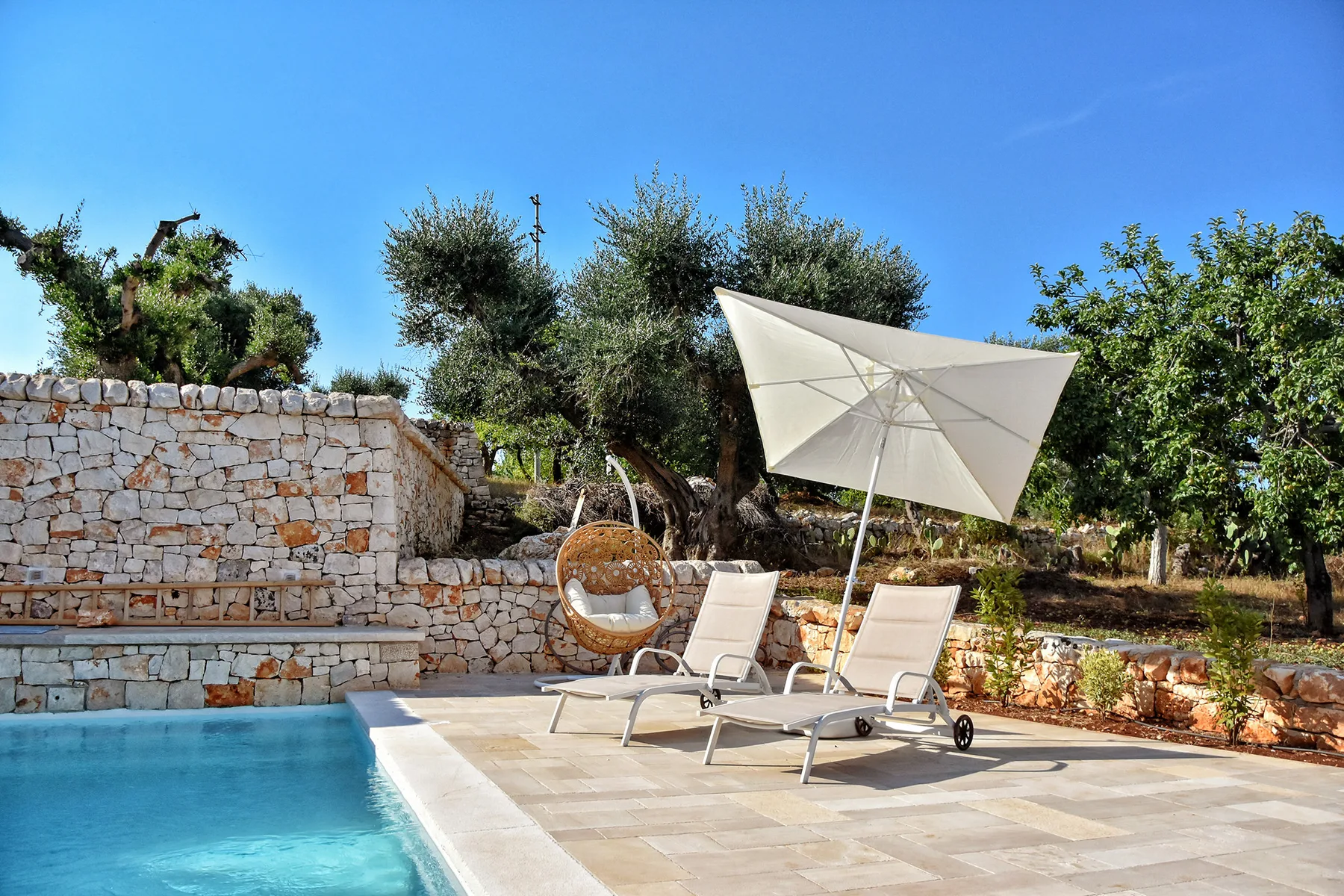 Una piscina al aire libre junto a una terraza de piedra con asientos reclinables y una sombrilla.