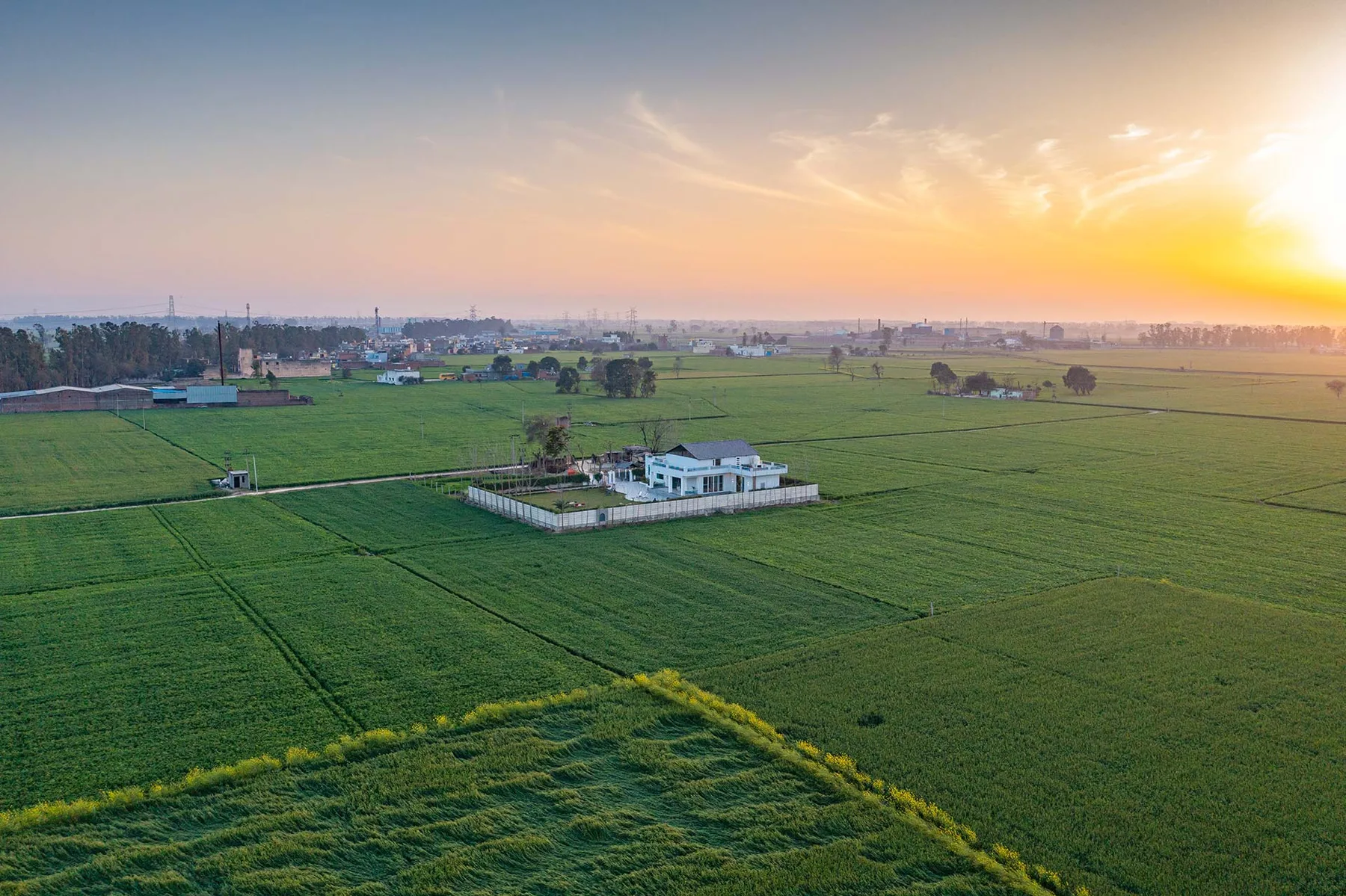 Vista aérea de campos verdes con un pequeño edificio blanco bajo el cielo al atardecer.