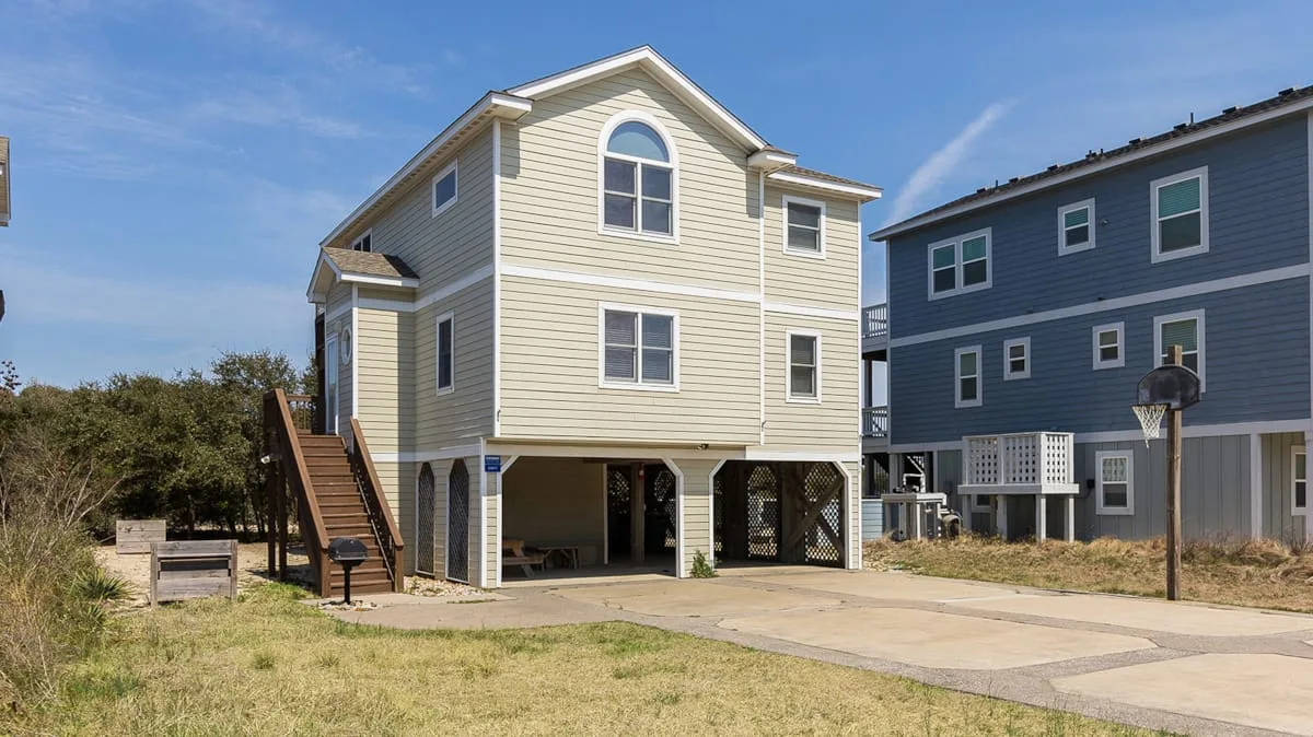 A raised coastal-style house stands beside a blue multi-story building.