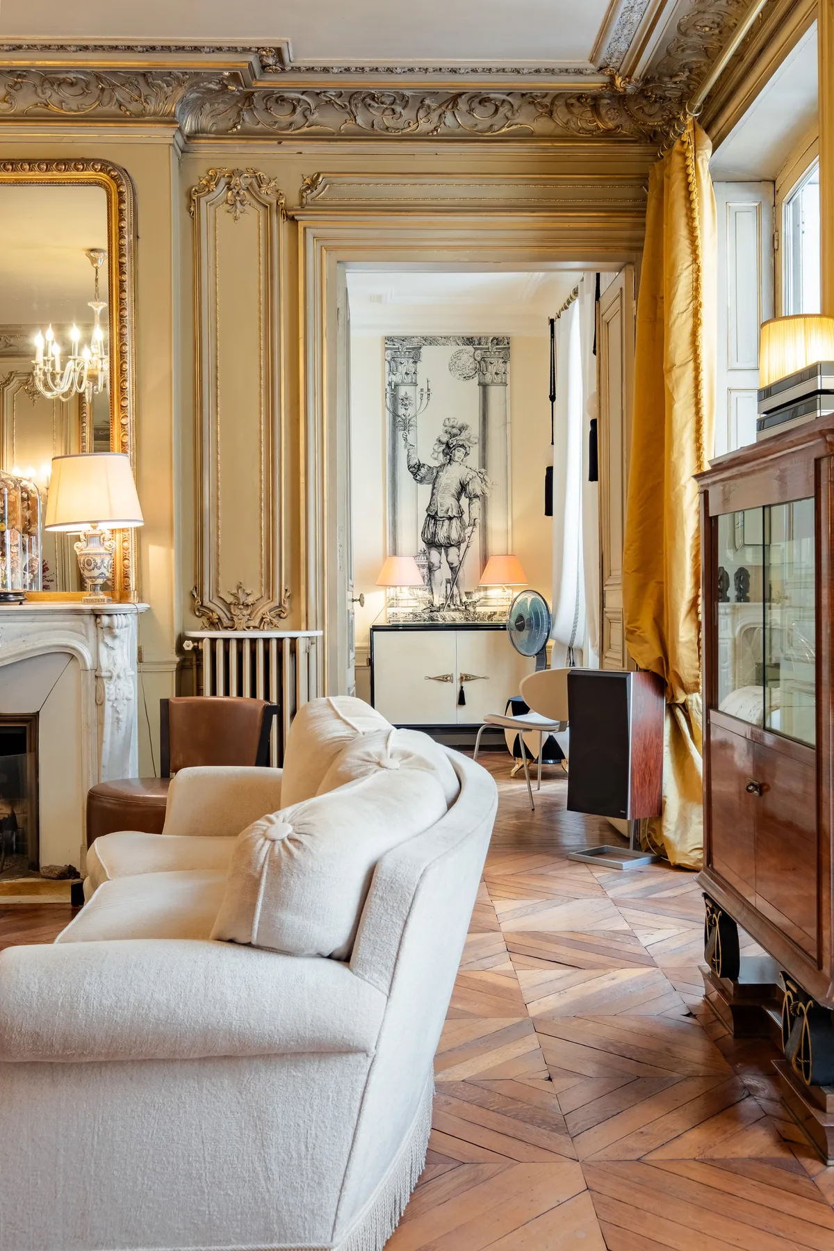An ornate living room with pale seating, decorative wall panels, and large windows.