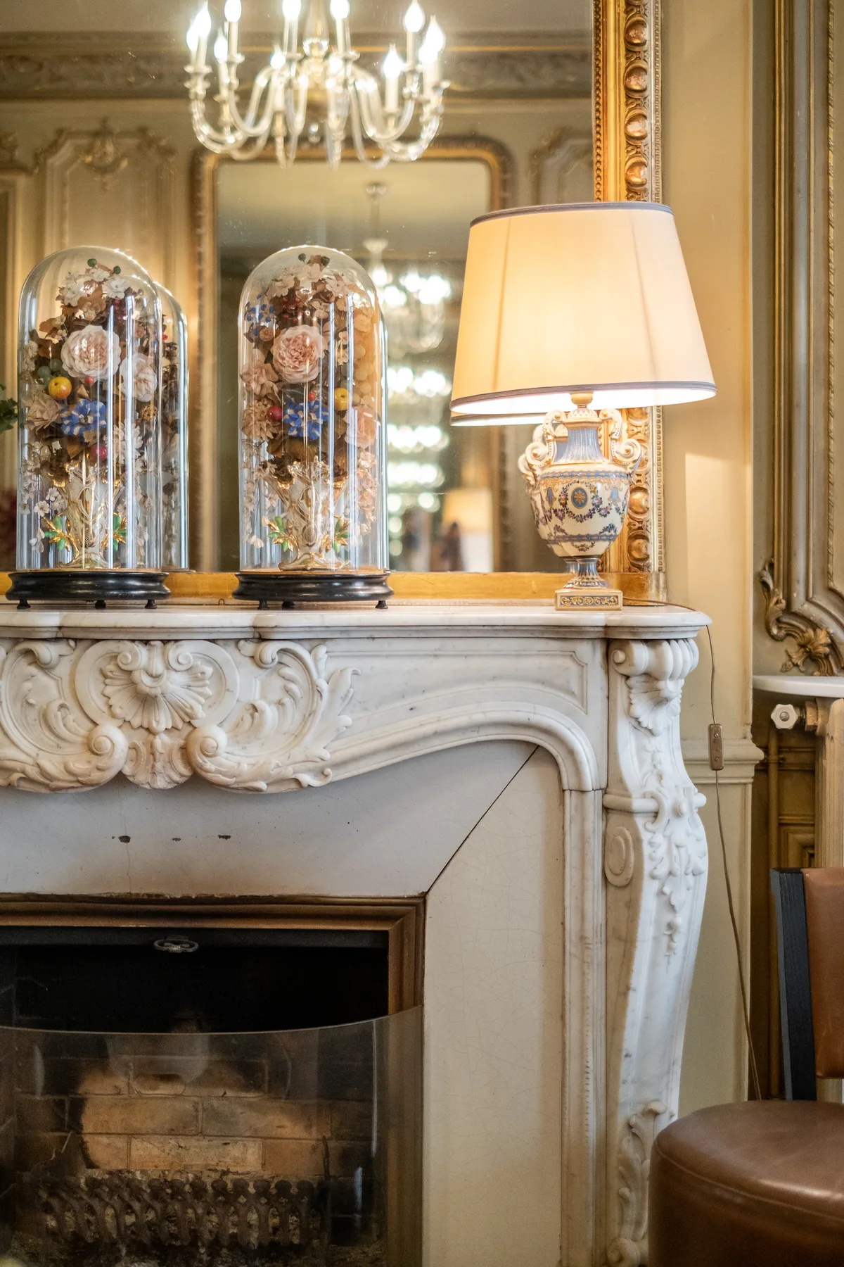 An ornate fireplace beneath a mirror, with decorative displays and a lit table lamp.