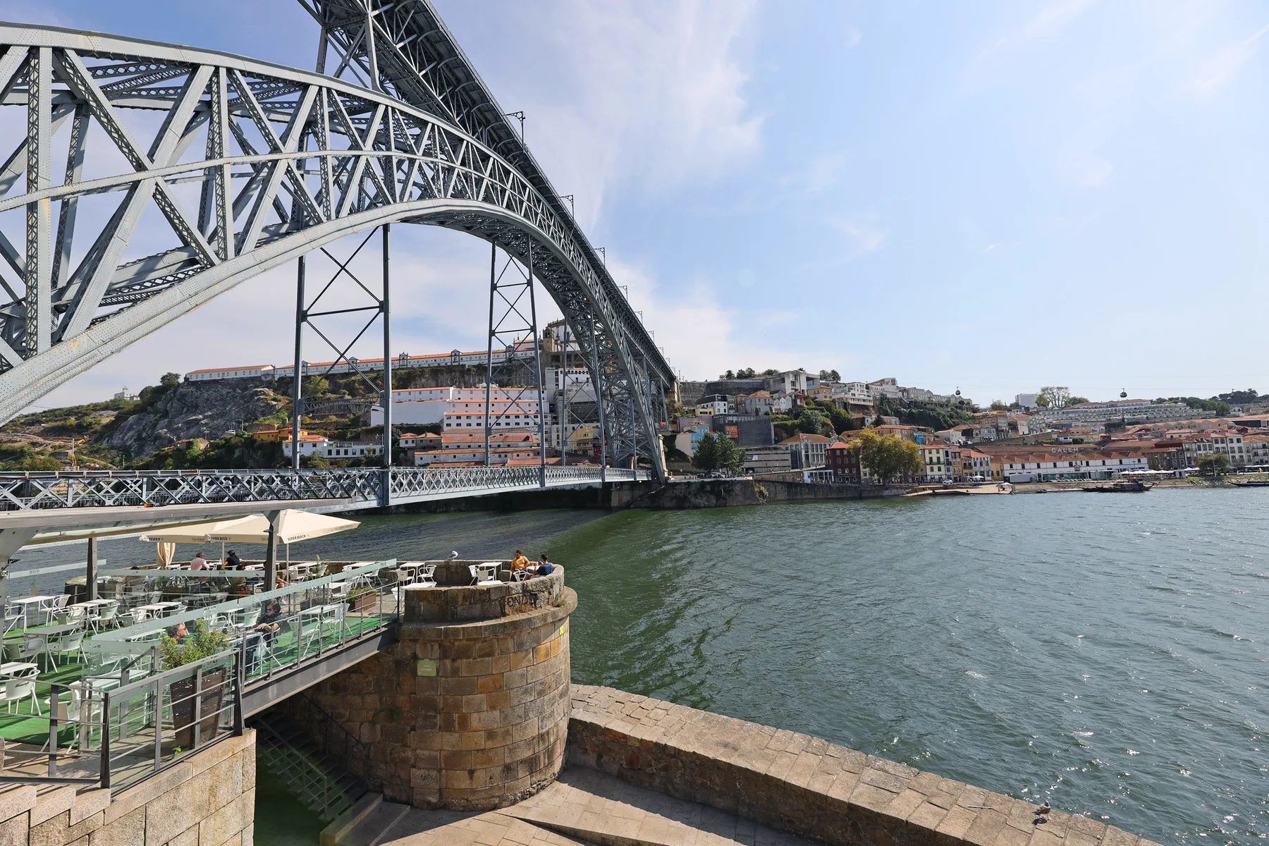 A steel arch bridge crosses the river beside a waterfront promenade.