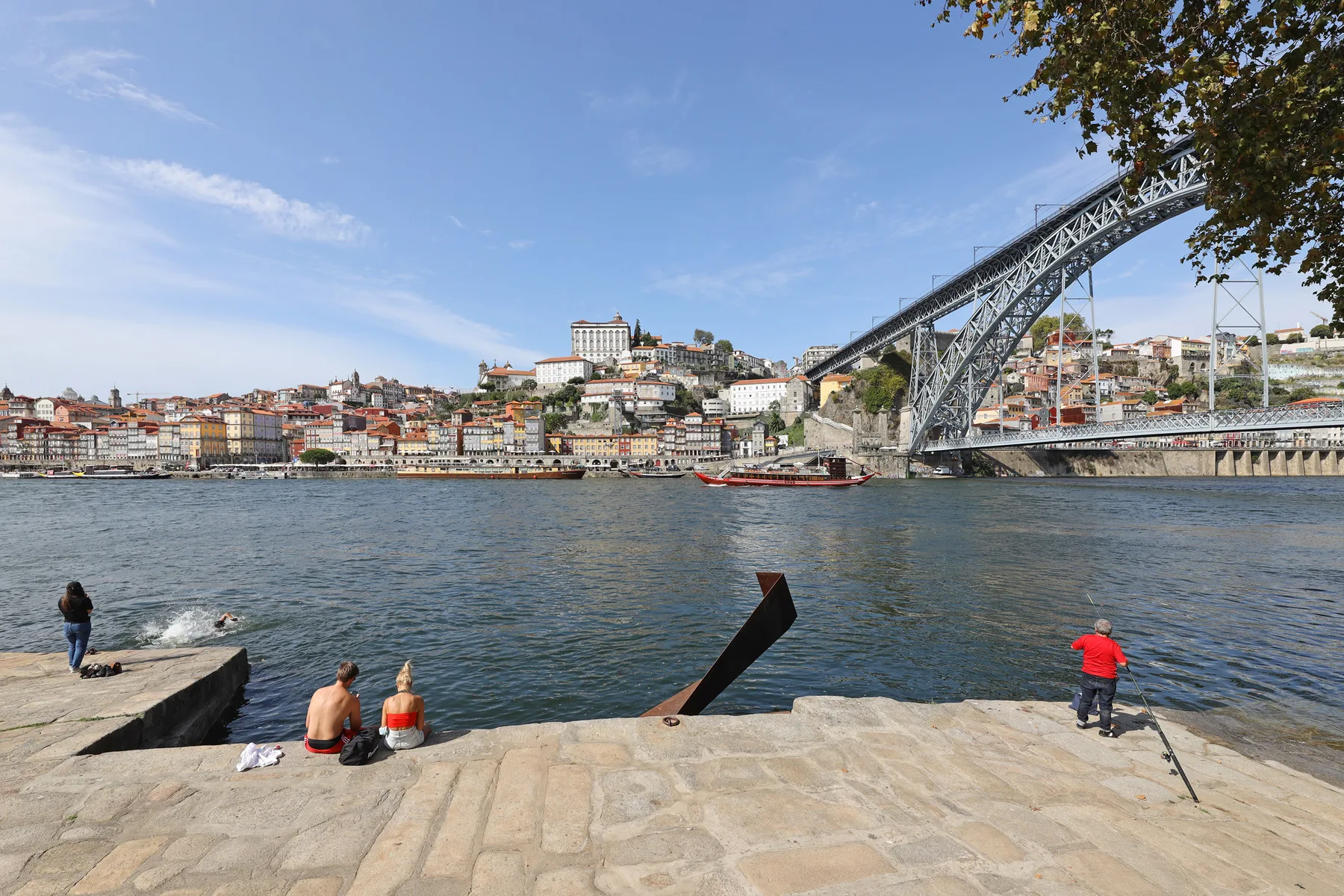 A riverside promenade overlooks the water, with a large arched bridge in the background.