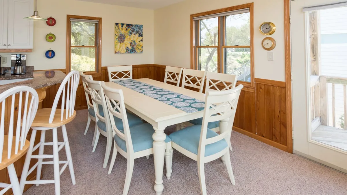 Un comedor con una mesa larga y asientos blancos junto a ventanas grandes.