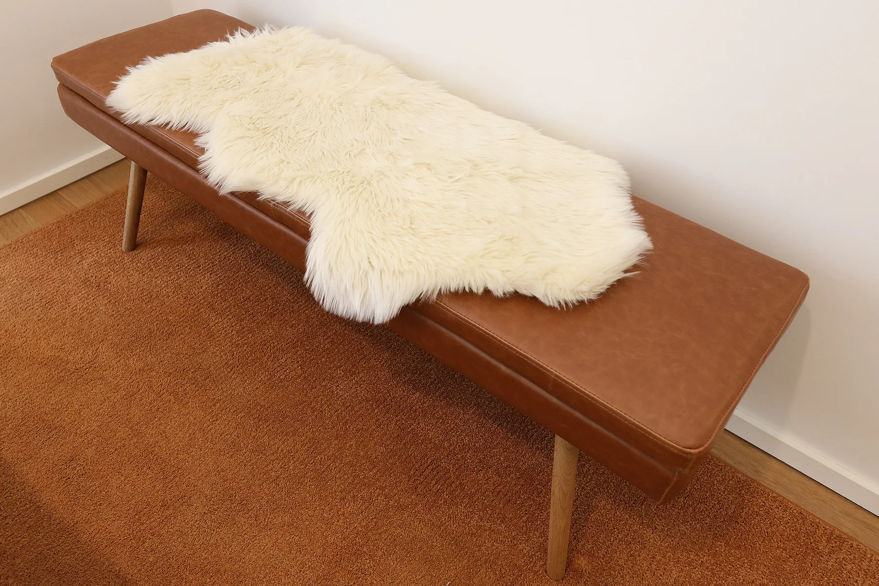 A brown upholstered bench with a white textured throw sits on a rust-colored rug.