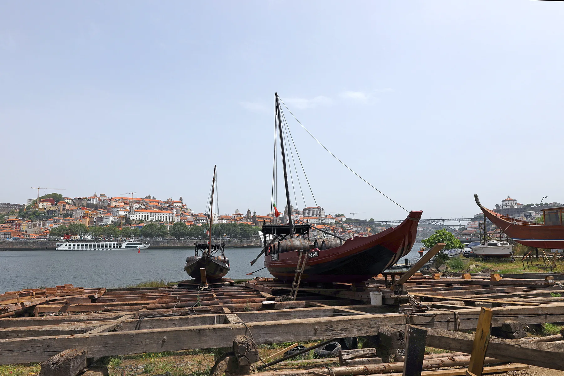 Historic boats rest on wooden supports beside the river, with hillside buildings across the water.